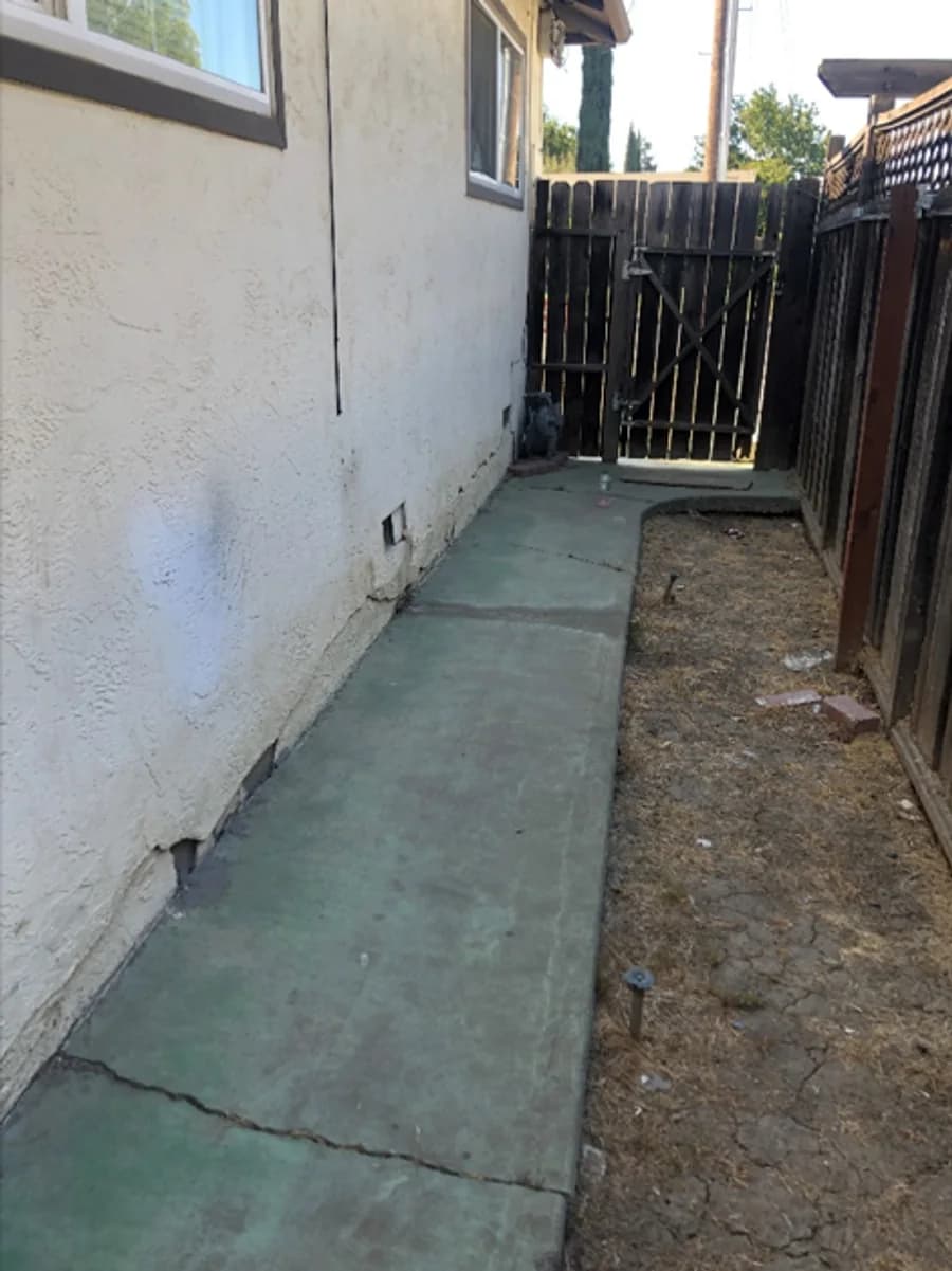 Narrow concrete walkway beside an exterior wall with wall and hardscape cracks near dry soil