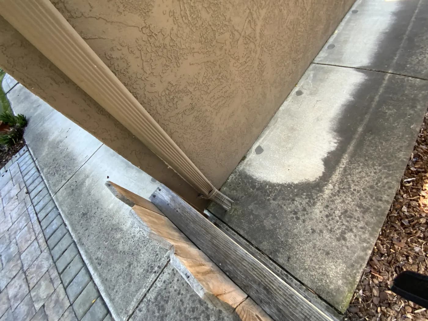 Downspout ending beside a low exterior corner with a dark wetting pattern across concrete hardscape